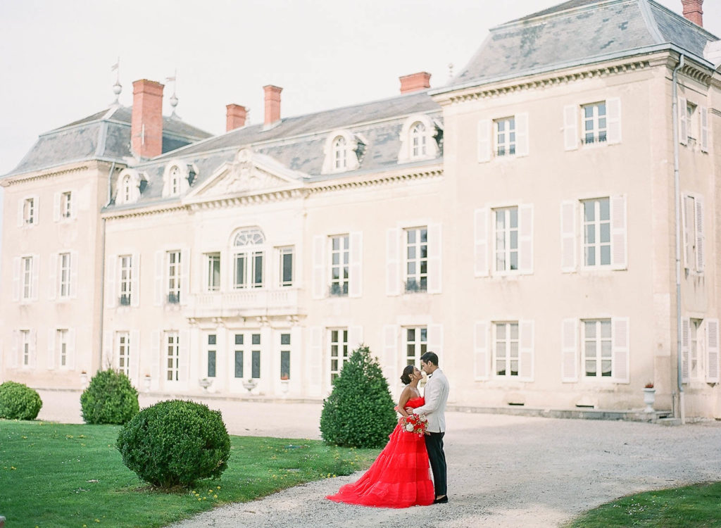 Bride in red monique lhuillier wedding dress and groom in a tux in front of chateau de varennes hugging and smiling